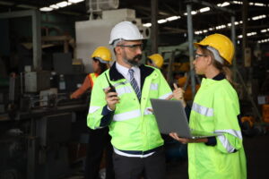 two workers wearing safety vests and hardhats have a conversation in a warehouse