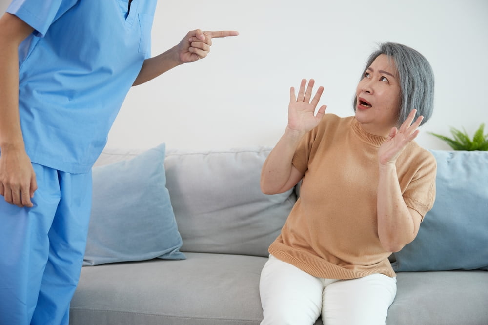 elderly woman sitting with a scared expression on the couch with hands up as she is menacingly pointed at by someone in scrubs