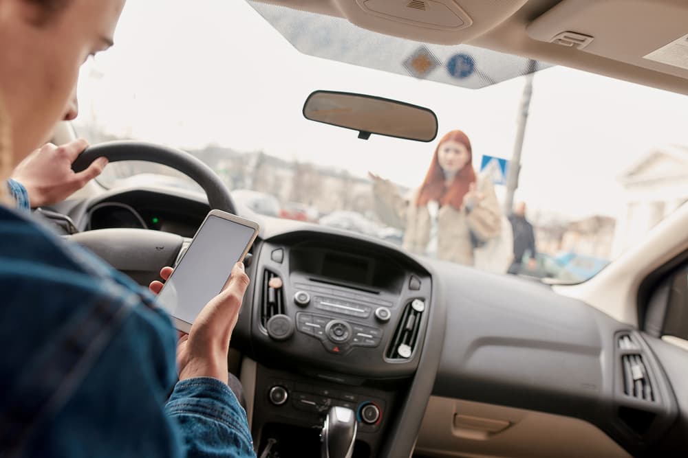 A man is checking his cell phone while driving, posing a dangerous distraction on the road, and is about to hit a female pedestrian.