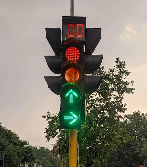 A traffic signal at an intersection is shown with a red light at the top, a yellow light in the middle, and two green arrow lights at the bottom. The green arrows indicate directions straight ahead and to the right. Above the lights, an electronic countdown display shows "00". The background includes trees and a cloudy sky.