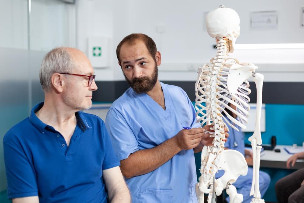 Chiropractic assistant with human skeleton explaining spine injury to retired patient.